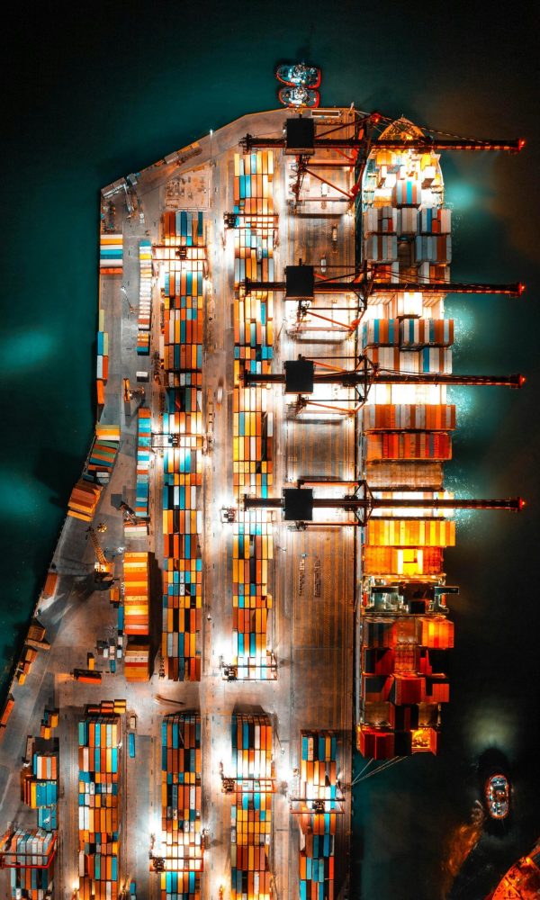Dramatic aerial view of a busy shipping port with colorful containers and bright lights at night.